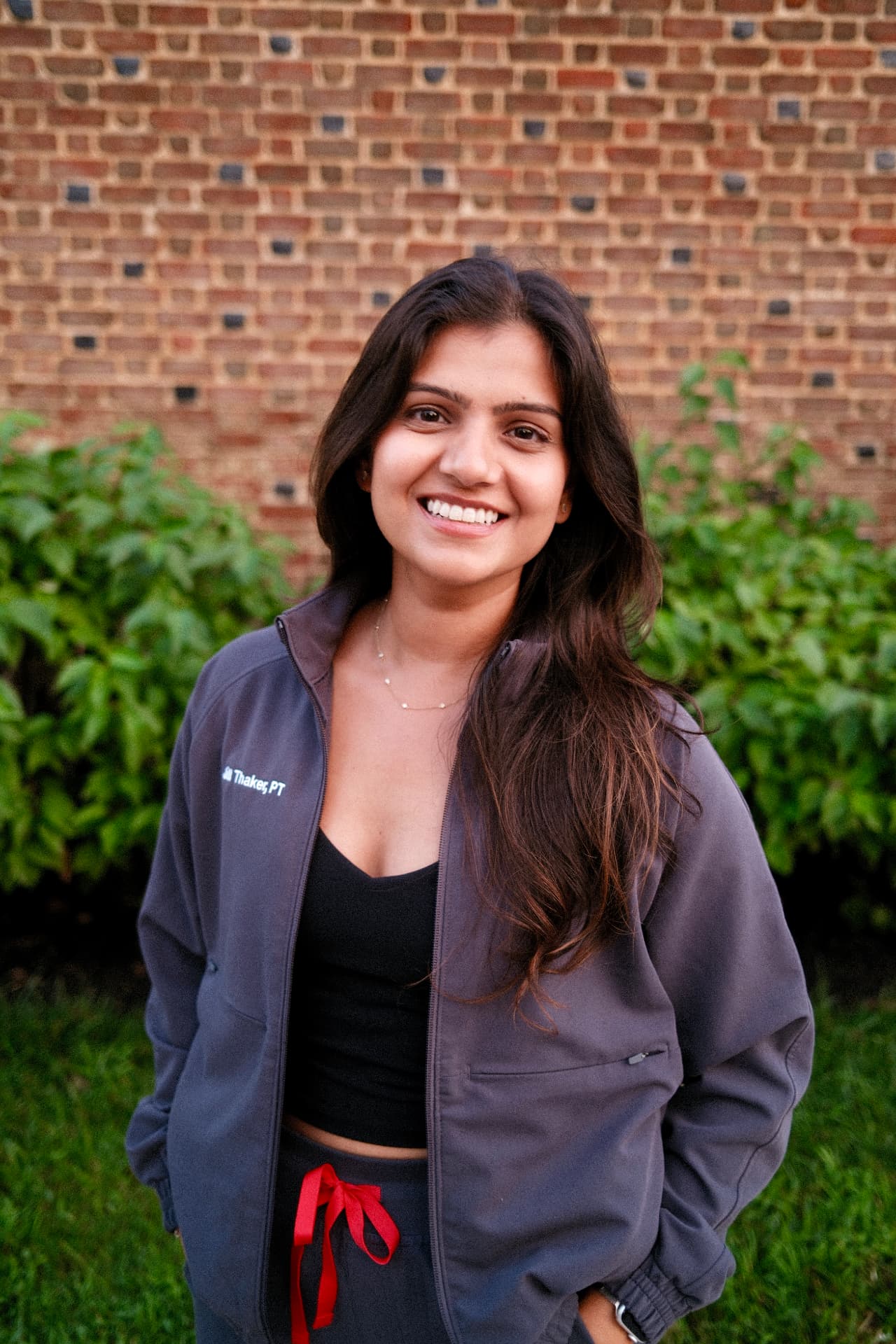 Dr. Jill Thaker in a charcoal physical therapy jacket, smiling in front of a brick wall