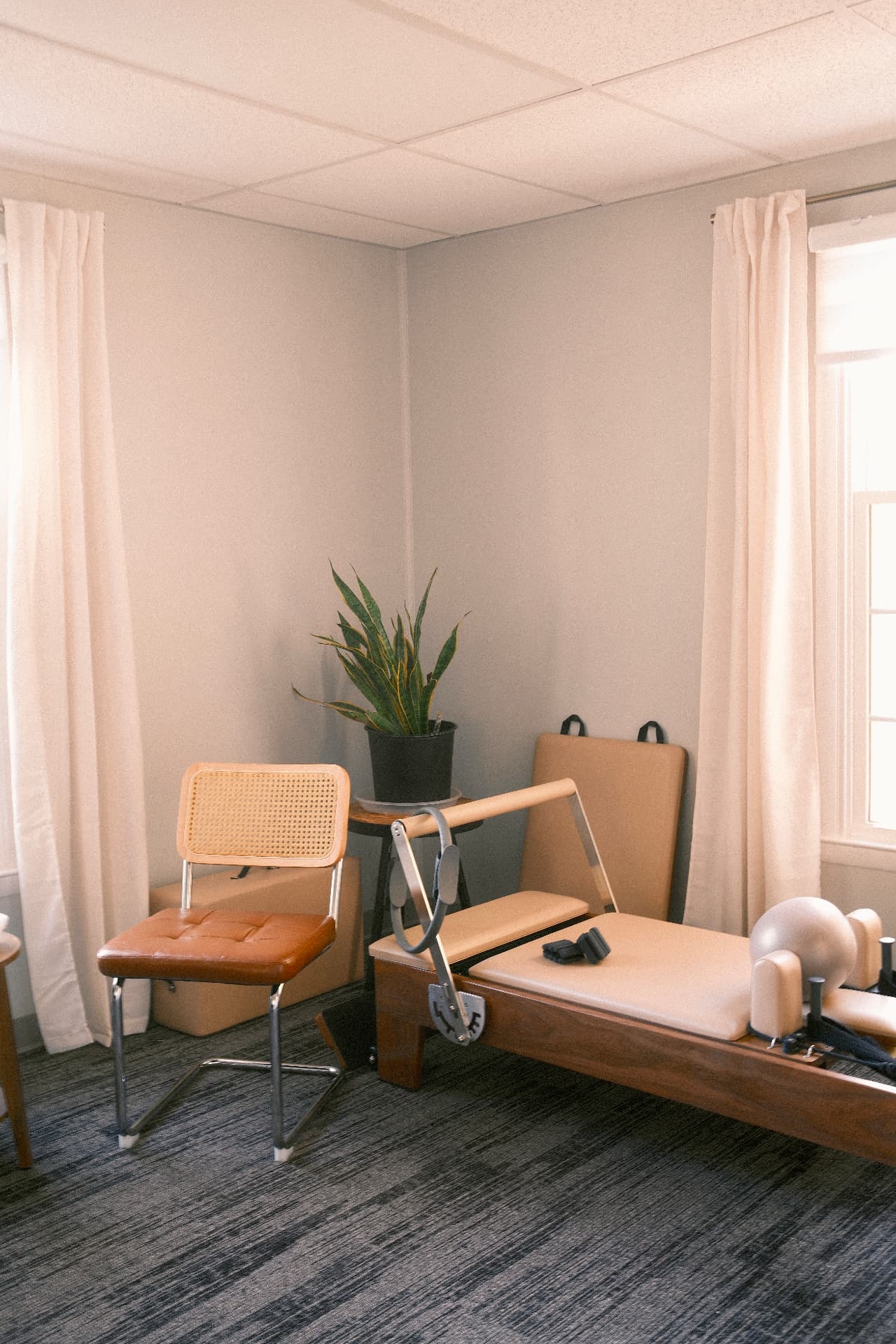 The walnut reformer in a sunlit corner of the studio, with a cane-backed chair, mats and a potted plant